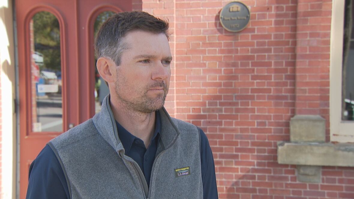 A man wearing a grey vest over a blue shirt stands in front of a red brick building