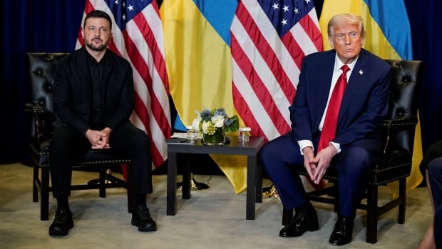 Two men sit in chairs with American and Ukrainian flags behind them.