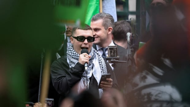 A person wearing sunglasses and a scarf holds a phone in one hand and a micophone in another as he speaks.