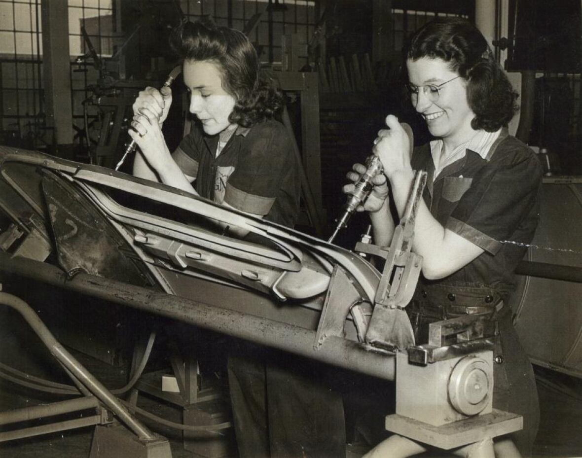 Two women factory workers working on a car part in a black and white photograph. It appears to be a historic photograph. 