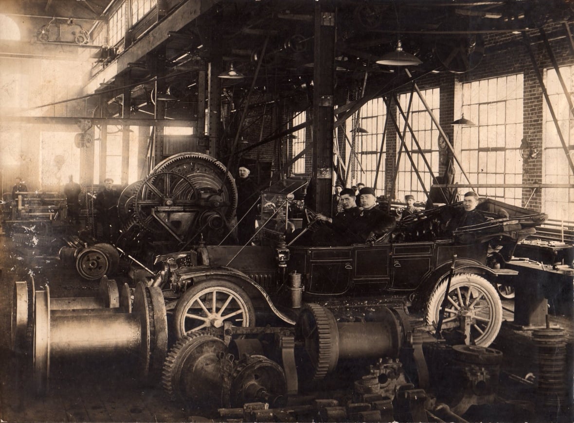 Black and white photo showing workers inside a car at a car factory. 