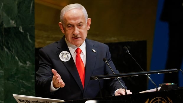 A person in a navy suit and red tie gestures while standing behind a podium.