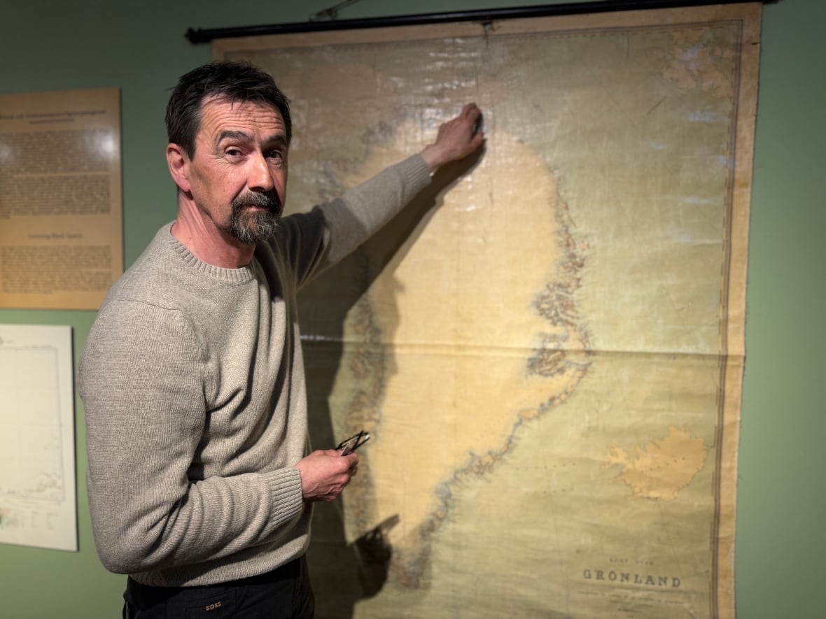 Um homem com camisa preta vestindo um suéter cinza aponta para um mapa da Groenlândia. O mapa está desbotado e tem uma tonalidade marrom.