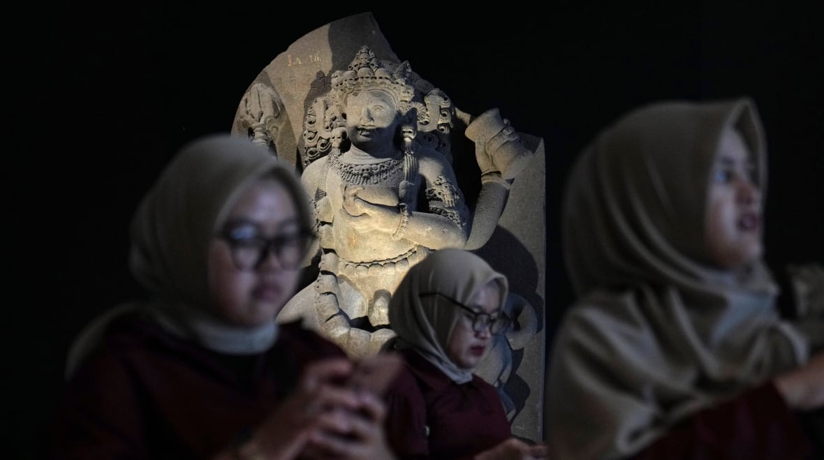 Três mulheres são visíveis no primeiro plano desfocado, com uma estátua de pedra ao fundo. A estátua está em foco e iluminada por um holofote.