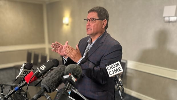 A man in a suit stands a podium surrounded by microphones and speaks to reporters in a hotel conference room.
