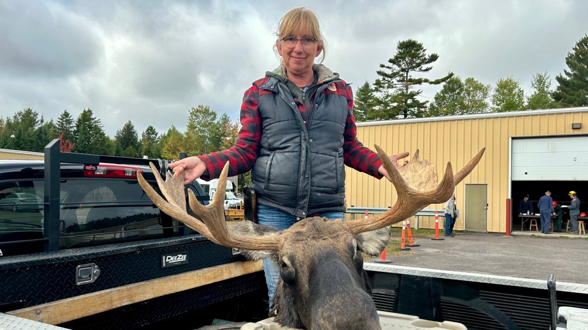 A woman with a red plaid sweater and blue vest poses in the back of a truck with the head of a moose. 