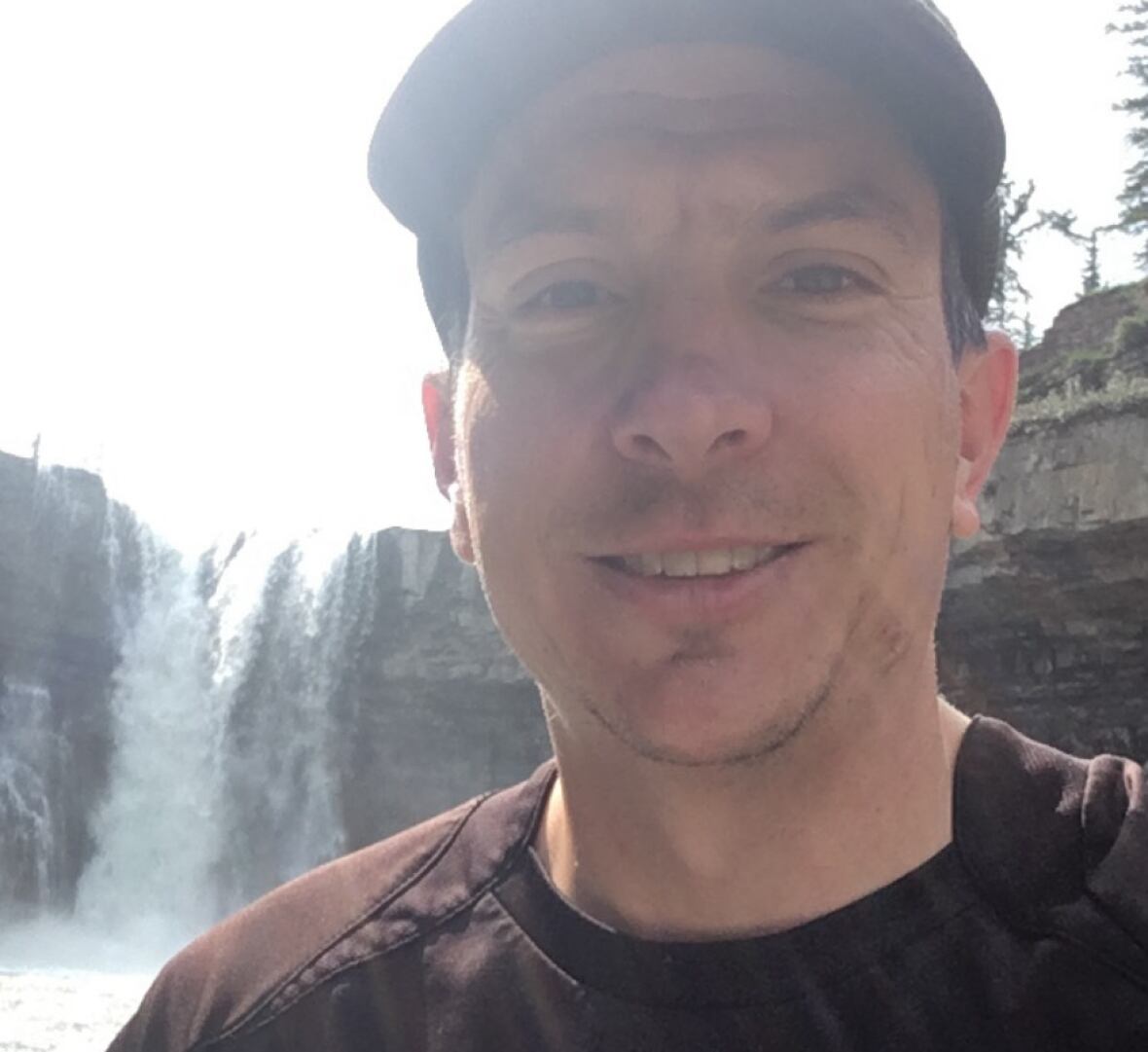 A man wearing a baseball hat poses for a selfie in front of a waterfall. 