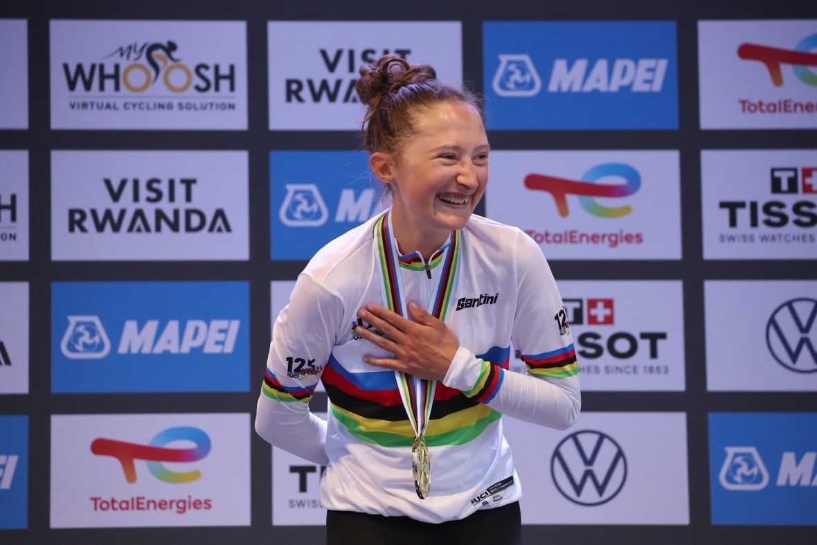 A female cyclist smiles while wearing a gold medal around her neck as she stands on a podium.