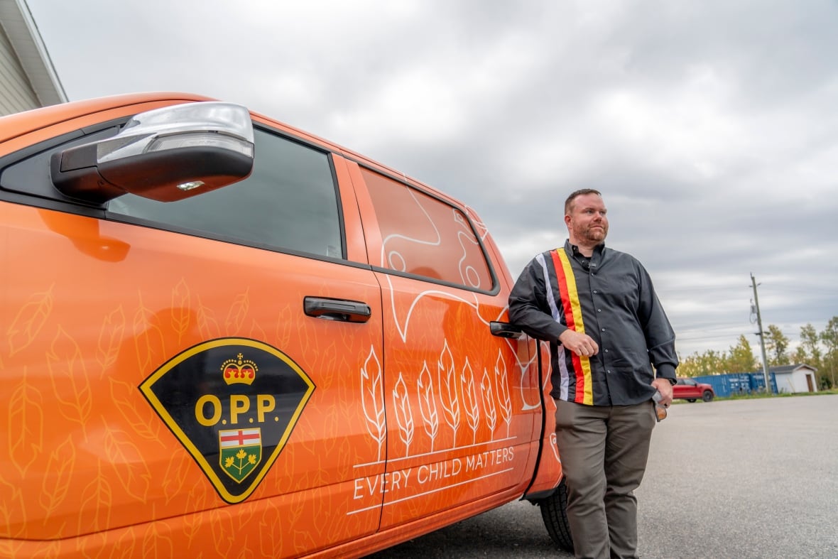 A person wearing a ribbon shirt is seen leaning against an orange police truck, which says "Every Child Matters."