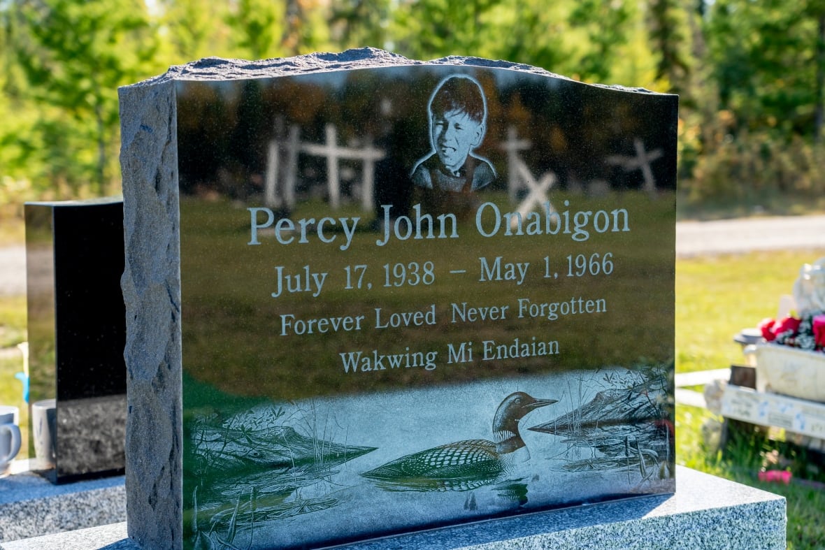A headstone is seen with the words "Percy John Onabigon, July 17, 1938 to May 1, 1966. Forever Loved, Never Forgotten. Wakwing Mi Endaian."