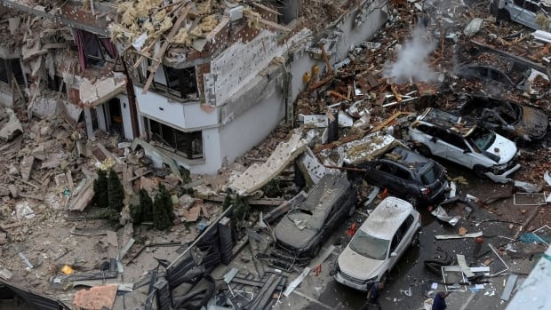 Destroyed buildings are seen from above as people cross a street filled with debris.