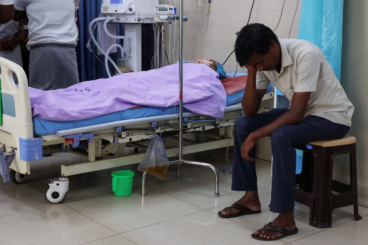 The patient lies in a rich hospital under a purple blanket. A man sits next to the patient with the head on hand.
