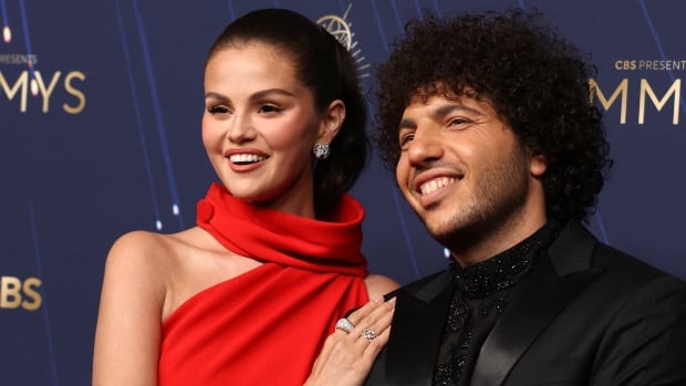 Selena Gomez, dressed in a red gown and sleeked back ponytail, rests her hand on Benny Blanco's shoulder while they pose for photos.