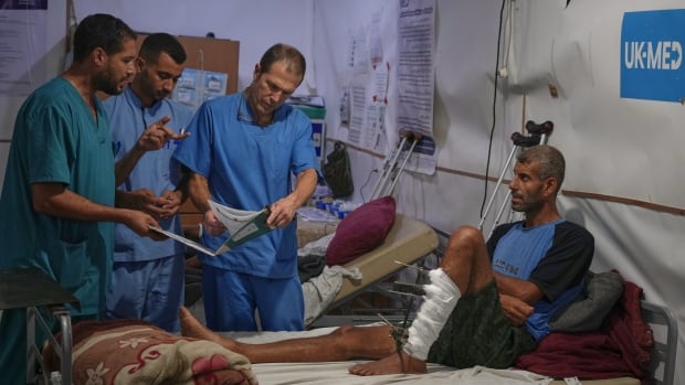 A man sits on a hospital bed with a bandaged leg, looking up at medical professionals.