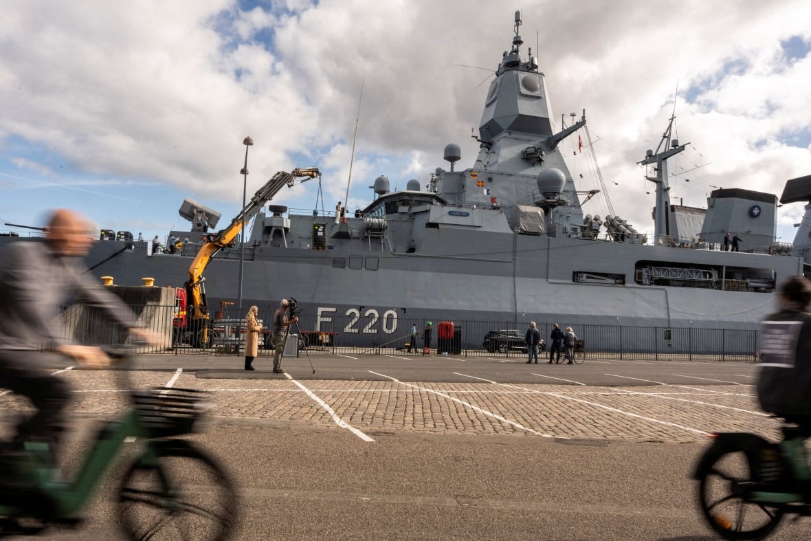 A large gray military boat is seen in an urban port, while pedestrians and cyclists pass.