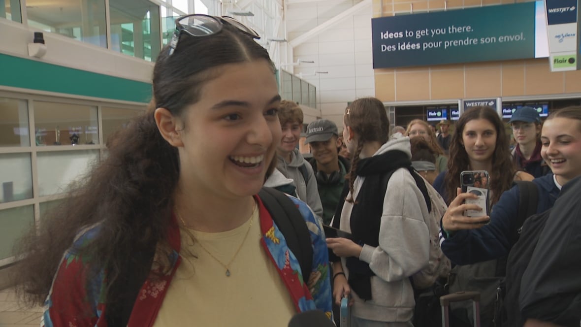 A young woman with long curly brown hair smiles at a crowded airport 