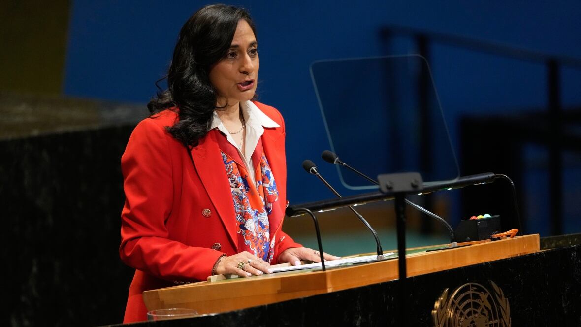 A woman in a red blazer speaks into a microphone at a podium.