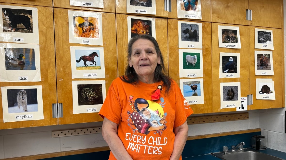 A woman wearing an orange "Every Child Matters" t-shirt stands in front of cupboards decorated with pictures of animals labeled in Cree.
