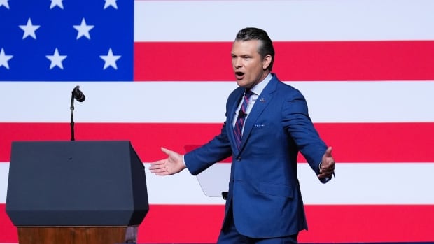 A man in a suit talks in front of a giant U.S. flag
