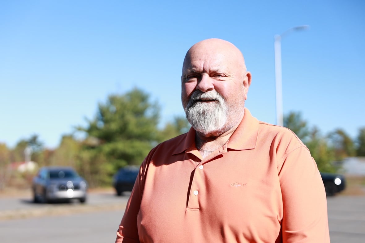 A man wearing an orange shirt stands for a portrait photo outside, and looks to the left.