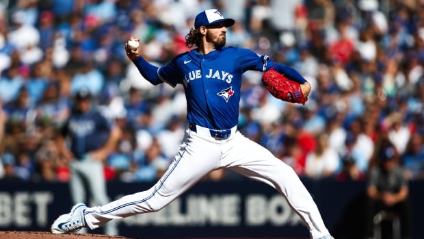 A male baseball pitcher for the Toronto Blue Jays winds up to throw the ball with his right hand as fans watch from the stands during a day game.