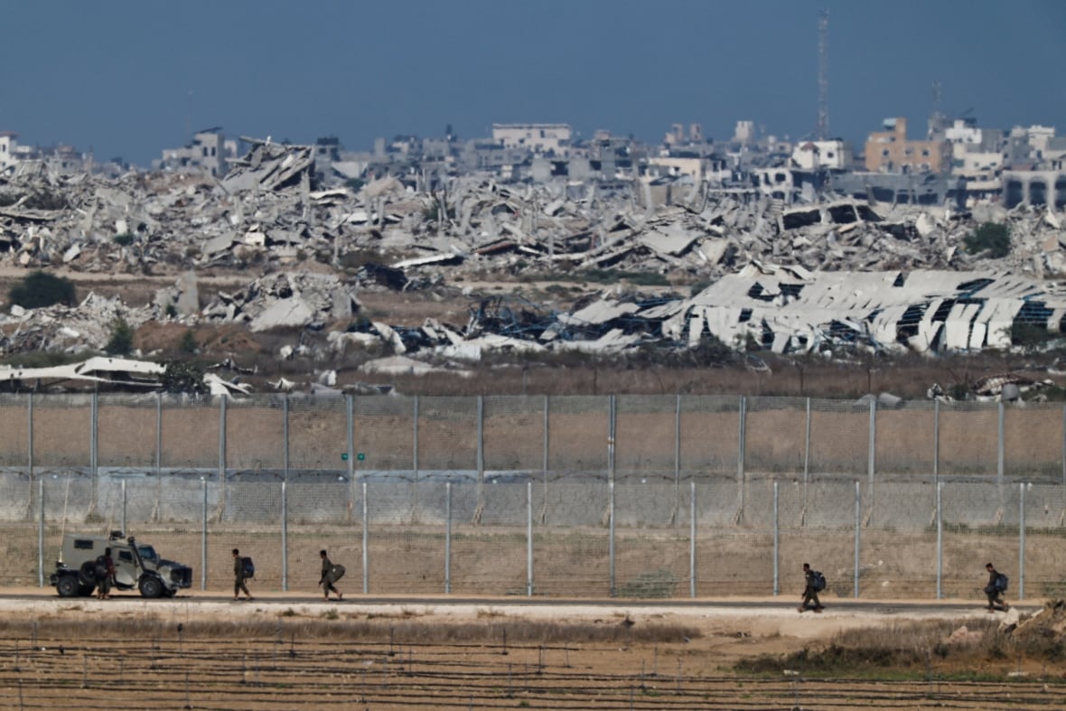 Soldiers walk on along a fence with destroyed buildings in the background.