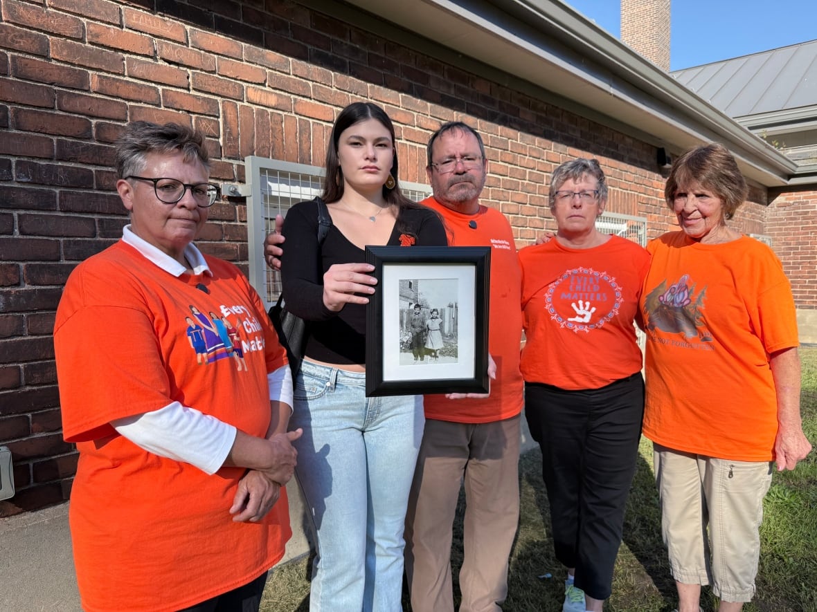 Five people, four in bright orange shirts, stand arm in arm. One holds out an old framed photo of two children.