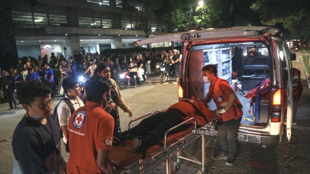 Paramedics wheel a stretcher, with a person laying on it, toward the back of an ambulance as other people stand on the street outside at night. 