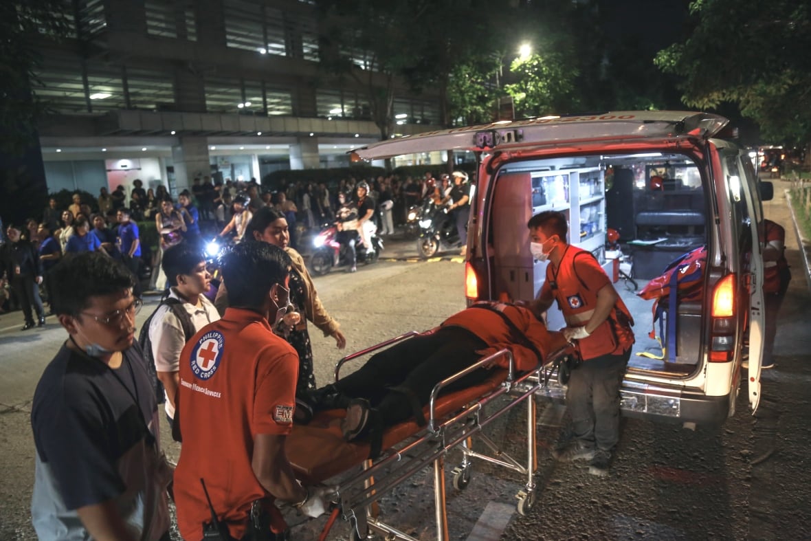 Paramedics wheel a stretcher, with a person laying on it, toward the back of an ambulance as other people stand on the street outside at night. 