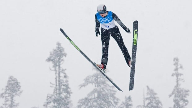 A male ski jumper flies in the air with legs extended and skis out in front of him.