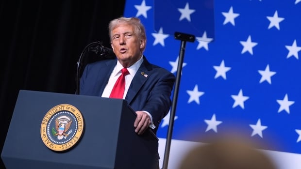 A man in a suit speaks into a microphone at a podium in front of an American flag.