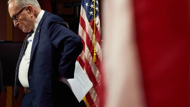 A profile view of a man in a blue suit looking downward with a sullen face as he stands with his hand on his lower back, holding papers, with a red, white and blue U.S. flag behind him. 