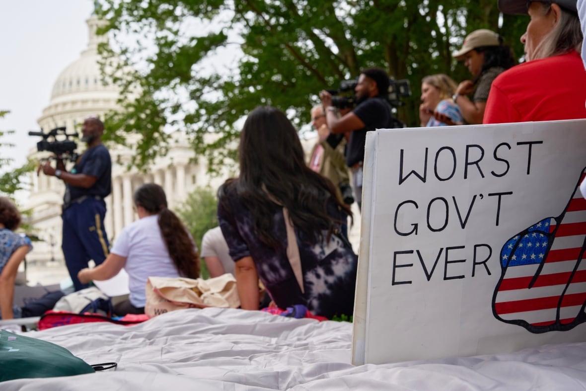 Reading a white banner "The worst government ever" He sits on a white blanket on the ground at the front of the image, in front of a crowd of people and a man speaking on a platform in front of a large white building with a long dome on top. 
