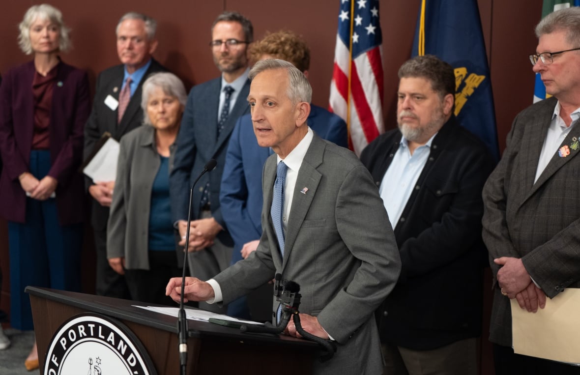 A group of men and women stand behind a man with gray hair wearing a gray suit, talking to a microphone.