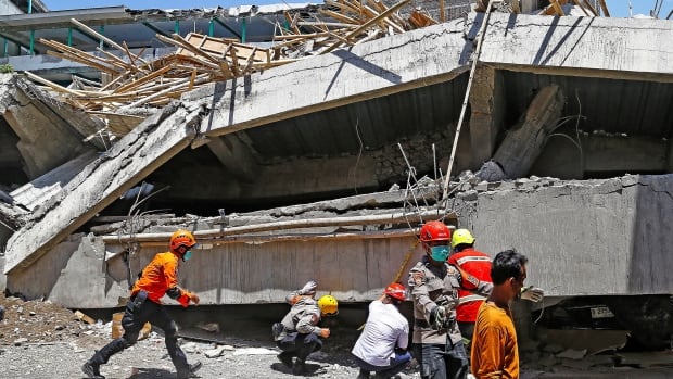 A half-dozen people, some in uniform and helmet are shown near a large piece of concrete that has collapsed to near the ground.