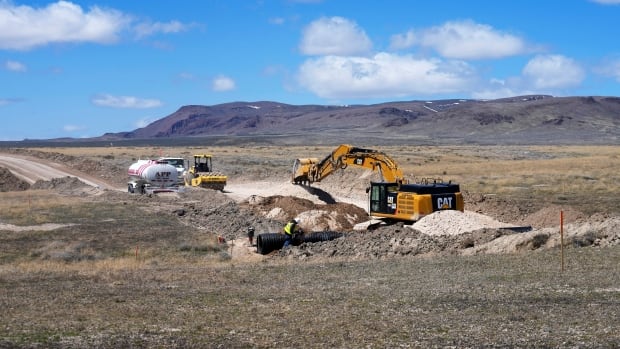an excavator digs into te ground on a construction site. other construction equipment is visible on a flat, mostly dirt stretch of land. in the background the sky is bright blue and a far-off hill or mountainous area is visible