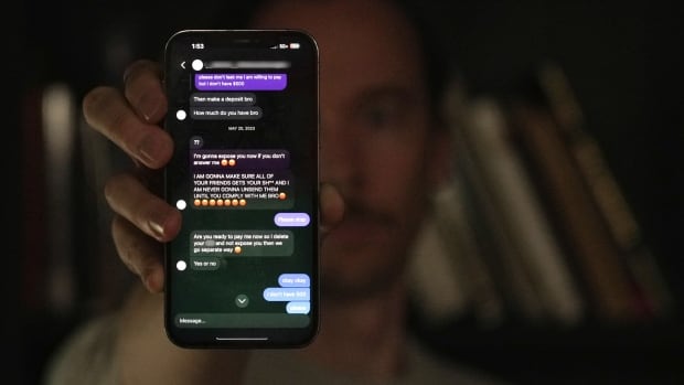 A man in a dark room holds up a phone right in front of him. The text on the phone are angry and demanding money.