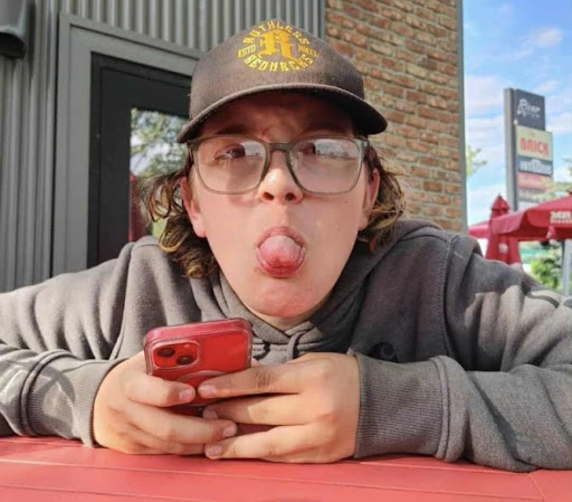 A boy sitting outside on a picnic seat with a phone in his hand. He holds his tongue with the camera.
