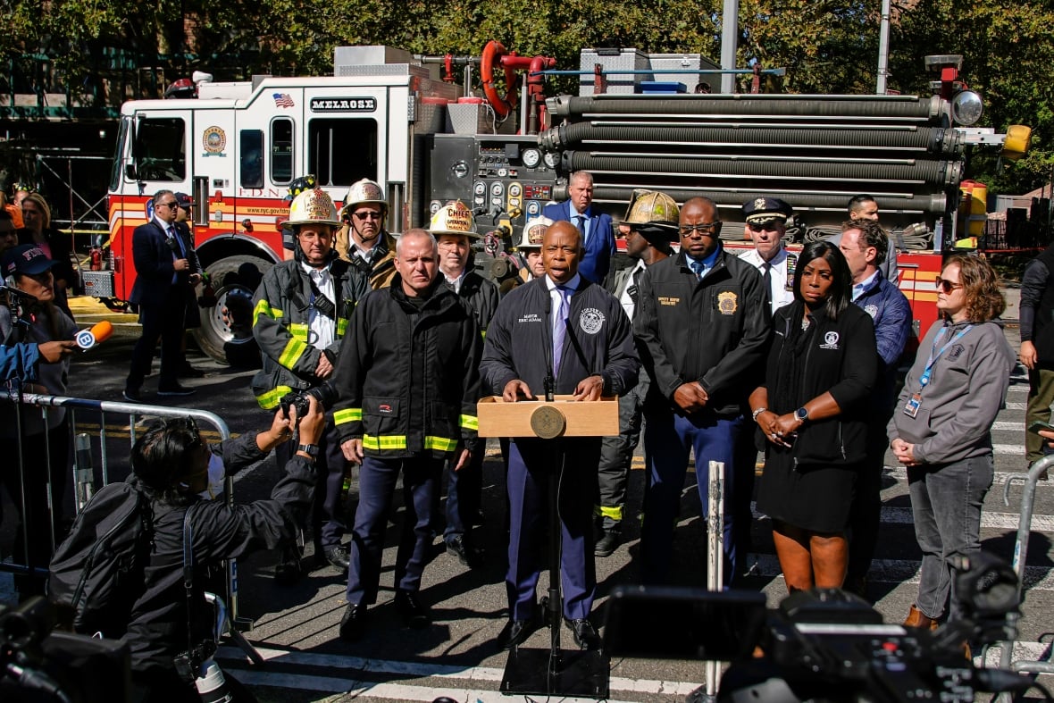 A man is seen speaking at a podium with a group of officials and a fire truck behind him.
