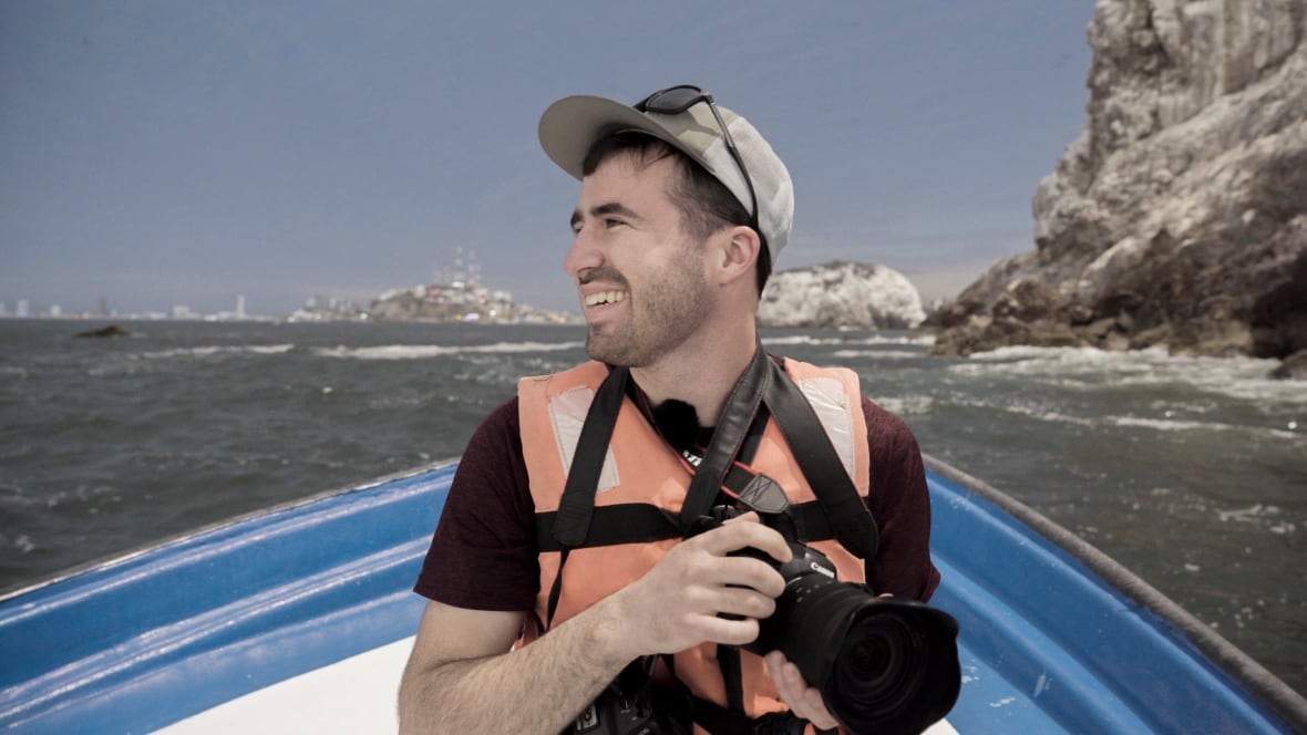 Um homem segurando uma câmera e sentado em um barco olha para o lado e sorri