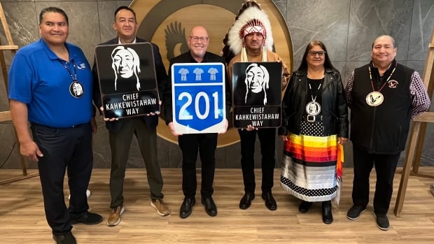 5 men and 1 woman stand side by side, one wearing a feathered headdress while holding a portrait sign, and two other men are holding highways signage on each side of him.