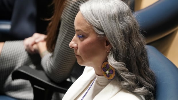 A woman with long grey hair and a white suit sits in a blue chair during a political event.