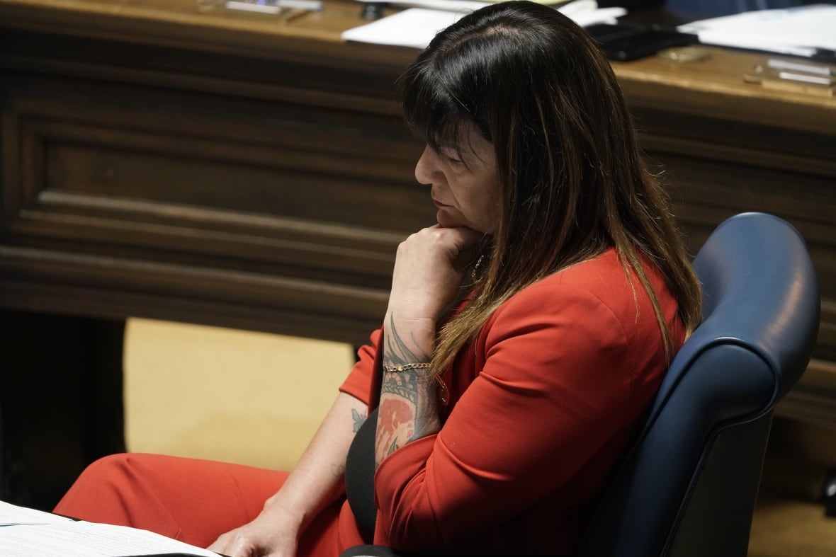 A woman in a red suit with long brown hair sits with her hand under her chin during a political event.