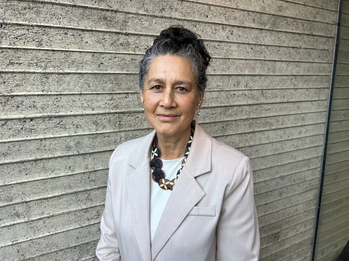 A woman wearing a cream-coloured coat stands in front of a striped wall.