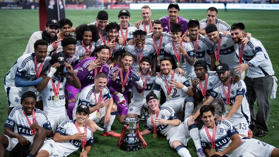 A group of soccer players wearing white uniforms pose on a field with a silver cup trophy.