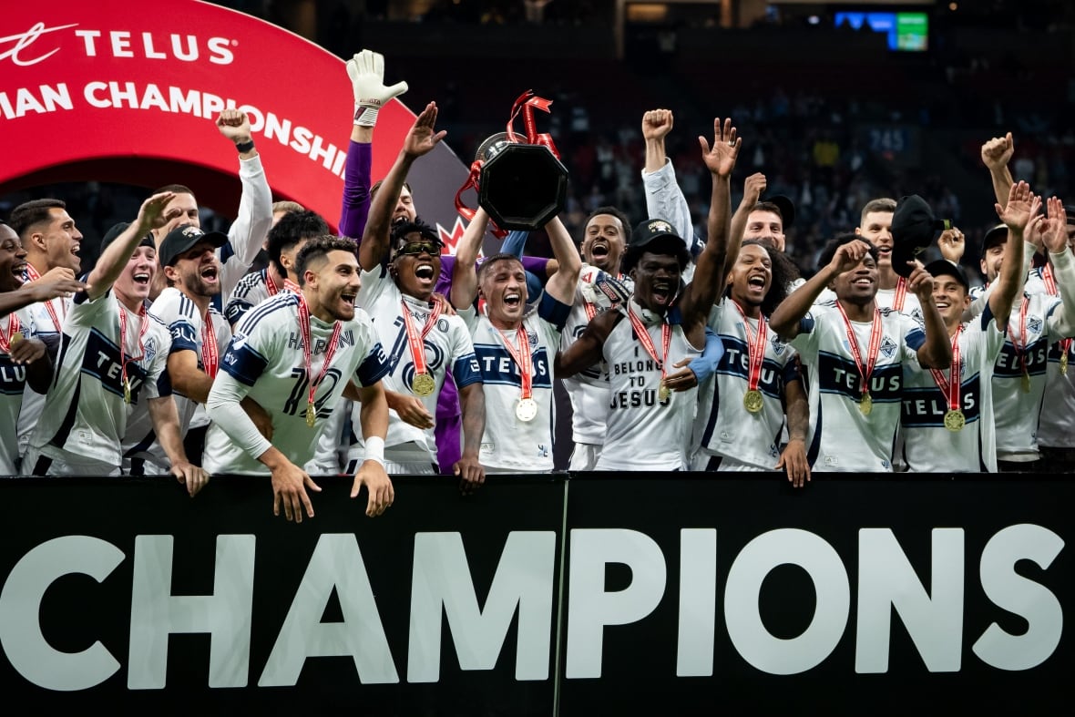 A group of soccer players in white uniforms stand behind a sign board and hoist a silver cup trophy.