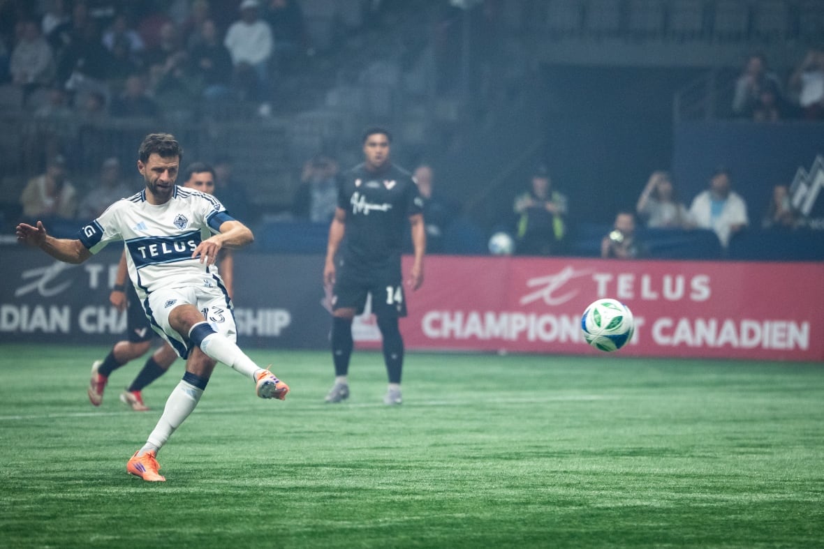 Thomas Müller, wearing a white and blue uniform, strikes a white, blue and green soccer ball from the penalty spot in the 18-yard box.