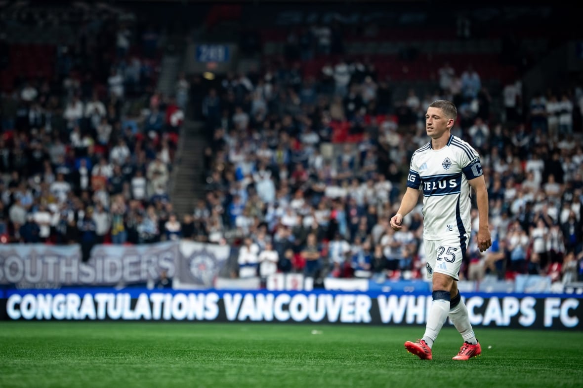 Ryan Gauld wearing orange football boots and a white and blue uniform walks on the pitch at B.C. Place. Fans are seen in the background.