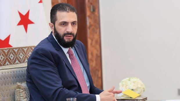 A man in a suit sits in front of a flag with red stars.