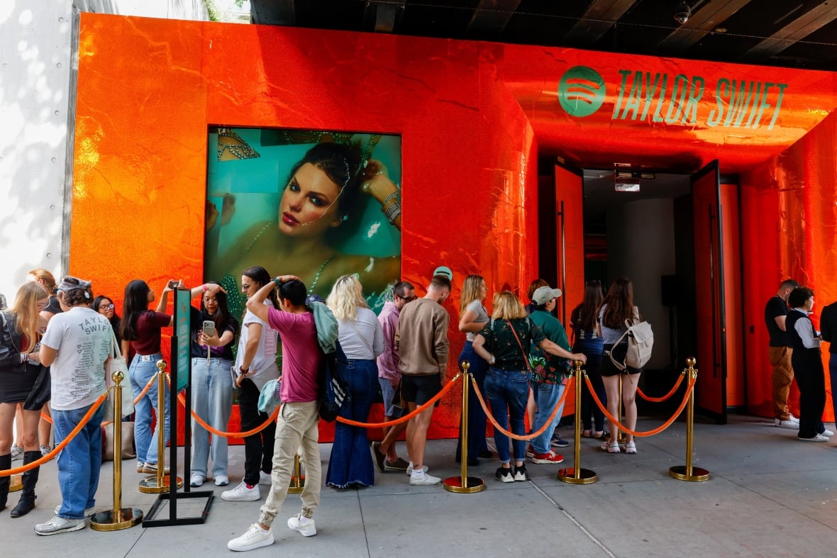 A group of people lined up on a sidewalk, behind a velvet rope, in front of a building with a bright orange facade, and the words 'Taylor Swift' over the entrance. On the wall above the lined up people is an image of an album cover with a woman, Taylor Swift, laying in water with her hand behind her head. 